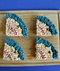 A cutting board with four shark heads on it. Each is a quarter of a circle with a blue strip on top and regular rice krispies treats filling in the rest with a white mouth and angry red squiggle and chocolate chip eye.