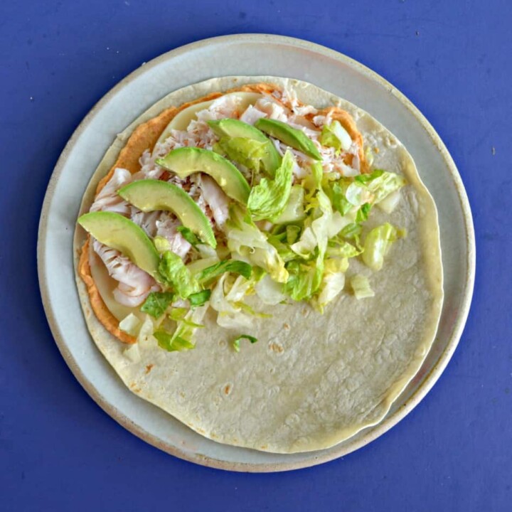 A plate with a wrap on it topped with hummus, turky, lettuce, and avocado slices.