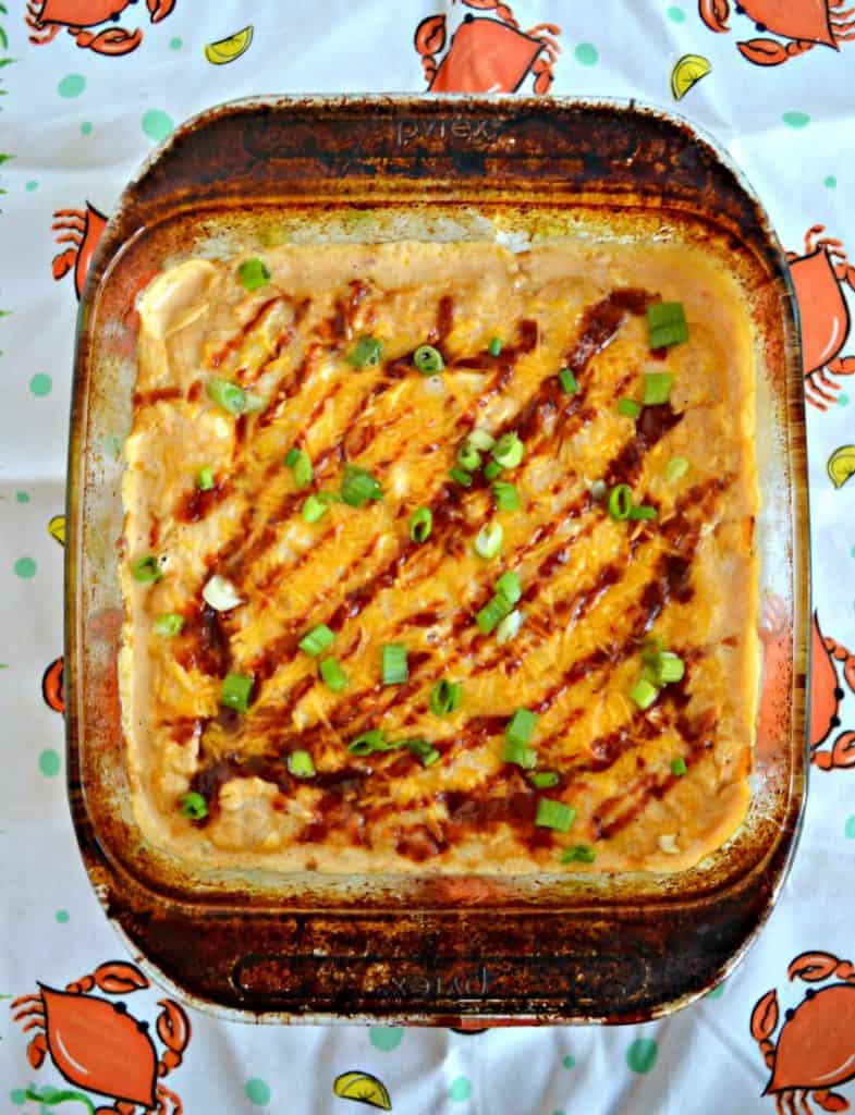 A baking dish filled with crab dip and topped with orange cheddar cheese, a drizzle of BBQ Sauce, and a sprinkle of green onions.