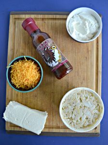 A cutting board with a block of cream cheese, a container of lump crab, a bowl of shredded cheese, a bowl of sour cream, and a bottle of BBQ sauce.