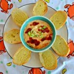 Plate with a small bowl in the middle filled with crab dip drizzled with BBQ sauce and sprinkled with green onions surrounded by buttered crostini on a crab tablecloth.