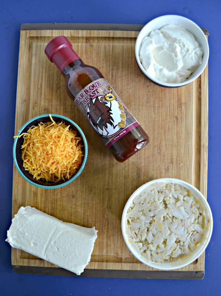 A cutting board with a block of cream cheese, a container of lump crab, a bowl of shredded cheese, a bowl of sour cream, and a bottle of BBQ sauce.