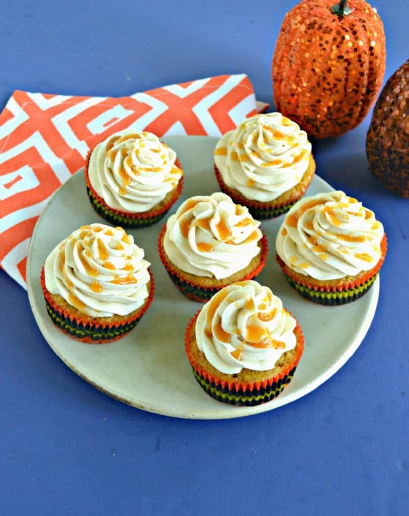A plate with 6 cupcakes drizzled with caramel on top of an orange and white napkin with 2 pumpkin behind the plate.