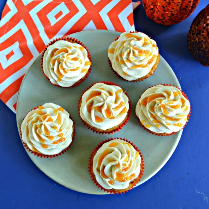 Top view of a plate with 6 cupcakes drizzled with caramel on top of an orange and white napkin with 2 pumpkin behind the plate.