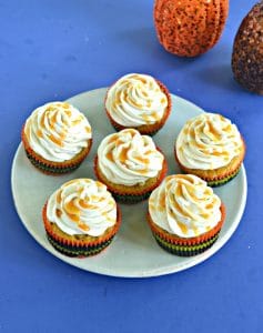 A plate of six butternut squash cupcakes topped with frosting and drizzled with caramel on a blue background with the bottom of a pumpkin showing in the corner.