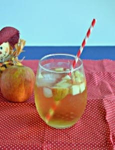 A stemless glass of Caramel Apple Sangria with apple pieces floating in it with a straw sticking out of it on a red background with a scarecrow holding and apple behind the beverage.