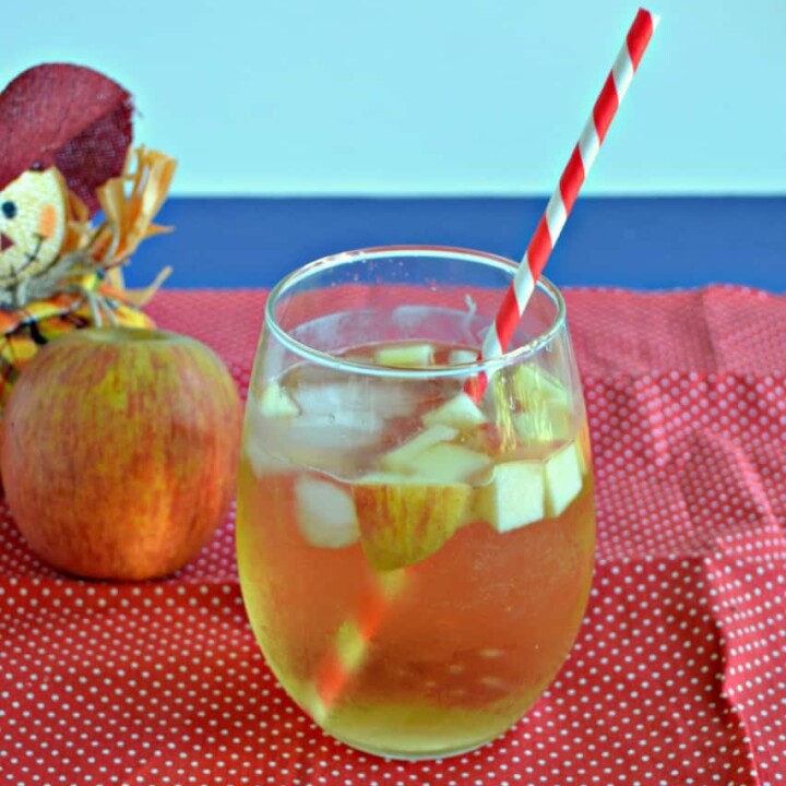 A stemless glass of Caramel Apple Sangria with apple pieces floating in it with a straw sticking out of it on a red background with a scarecrow holding and apple behind the beverage.
