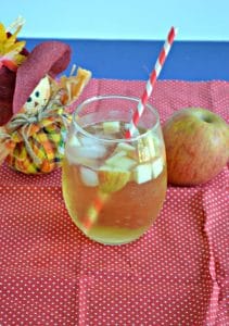 Close up view of a stemless glass of Caramel Apple Sangria with apple pieces floating in it with a straw sticking out of it on a red background with a scarecrow holding and apple behind the beverage.