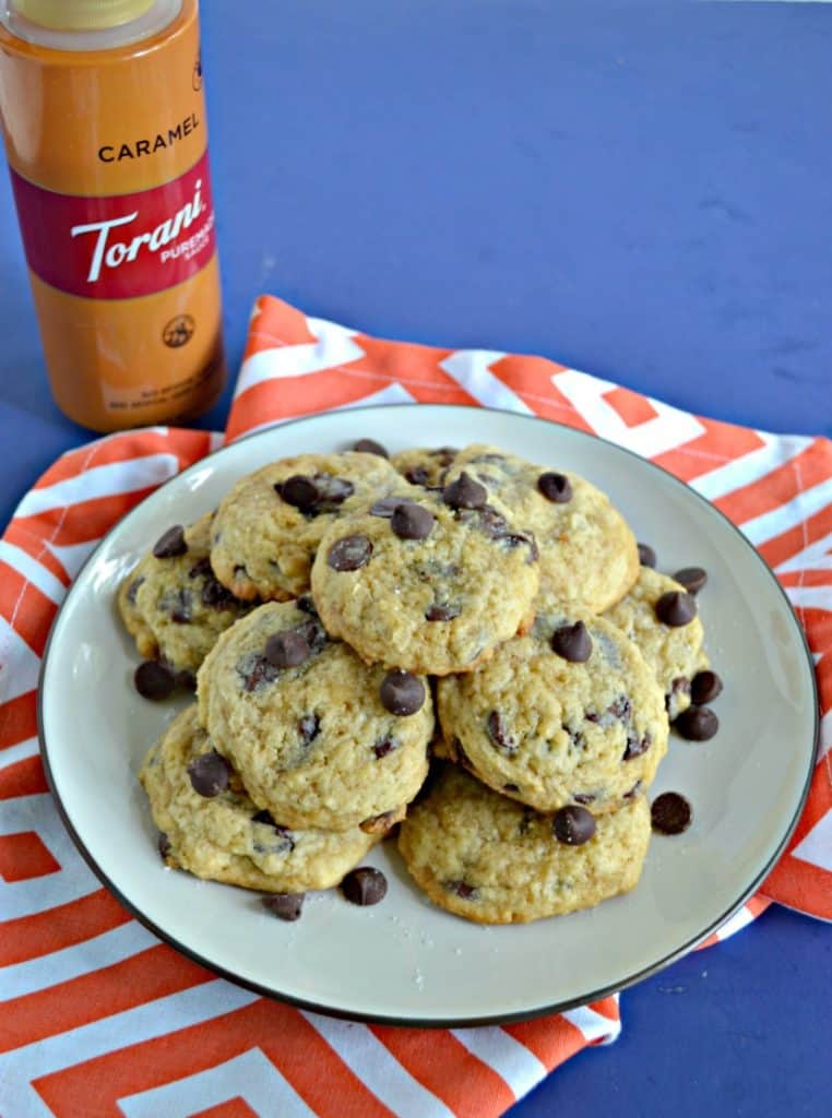 A plate stacked high with chocolate chip cookies set on an orange napkin with a bottle of caramel sauce on the left side.
