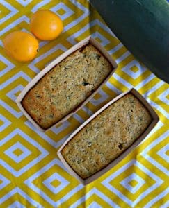 A yellow and white placemat with two mini loaves of bread sitting diagnal with two yellow lemons in one corner and a green zucchini in the other corner.