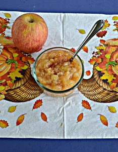 A bowl of apple and pear sauce with a spoon sticking out of it with an apple behind the bowl sitting on a cornucopia background.