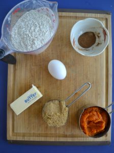 A cutting board with a cup of flour, a small bowl with a spice mixture, one egg, half a stick of butter, a half cup of brown sugar, and a half cup of pumpkin sitting on top of it.