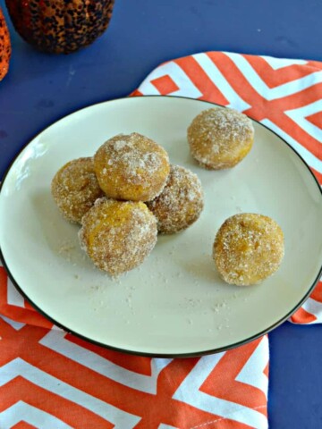 A plate with three donut holes piled with one more on top along with two donut holes on the opposite side of the plate sitting on an orange and white napkin with 2 pumpkins barely showing in the top left hand corner.