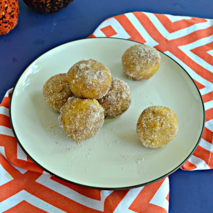 A plate with three donut holes piled with one more on top along with two donut holes on the opposite side of the plate sitting on an orange and white napkin with 2 pumpkins barely showing in the top left hand corner.