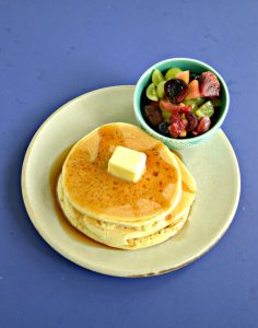 A front view of a plate piled high with 3 pancakes topped with syrup and a pat of butter, a bowl of fresh fruit on the side, all on a blue background.