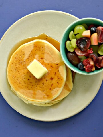 A plate piled high with 3 pancakes topped with syrup and a pat of butter, a bowl of fresh fruit on the side, all on a blue background.