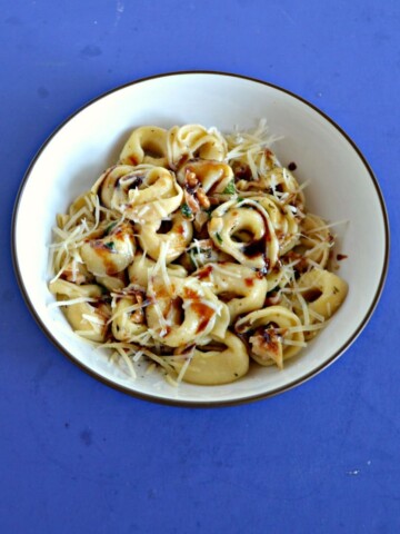 A bowl of tortellini topped with parmesan cheese and a balsamic drizzle on a blue background.