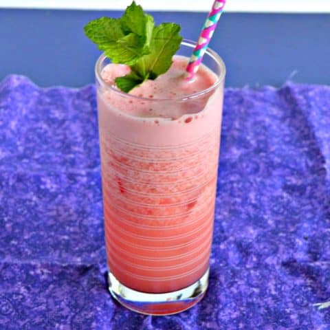 A bright pink cocktail that is frothy on the top with a pink and blue straw coming out of it and a bright green sprig on mint on top on a purple napkin.