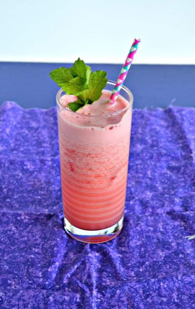 A bright pink cocktail that is frothy on the top with a pink and blue straw coming out of it and a bright green sprig on mint on top on a purple napkin.