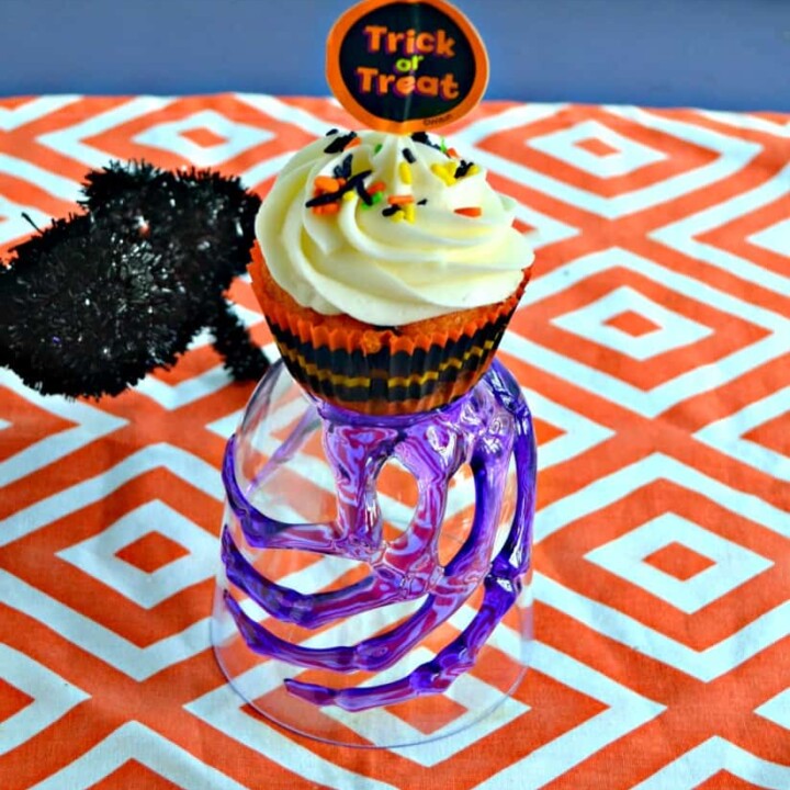 A glass with a purple skeleton hand holding a cupcake with white frosting and a trick or treat topper on top of an orange and white background with a black spider climbing up to the cupcake.