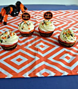 Four cupcakes with white frosting and black and orange sprinkles with a pumpkin topper on an orange and white background with an orange and black spider creeping up on them from the back left.