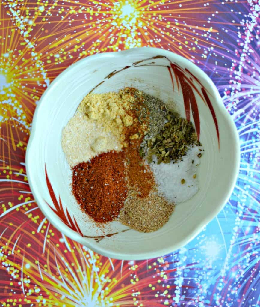 A white bowl filled with a teaspoon of red, brown, white, black, and yellow spices on a red, orange, blue, and purple fireworks background.