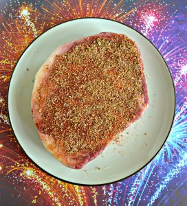 A huge ribeye covered with a rub on a white plate sitting on a red, orange, blue, and purple fireworks background.