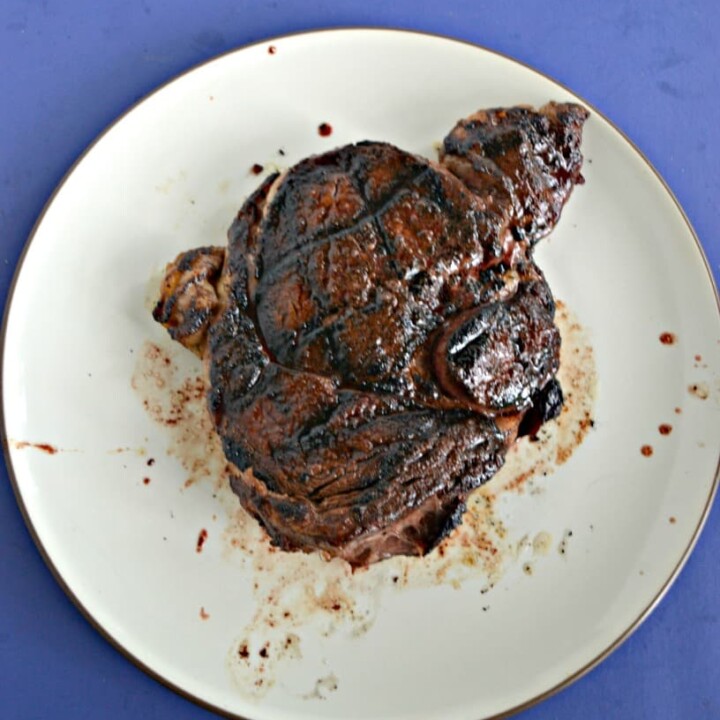 A large steak with grill marks cooked to dark brown sitting on a white plate on a blue background.