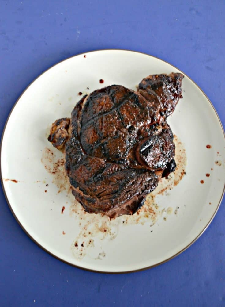 A large steak with grill marks cooked to dark brown sitting on a white plate on a blue background.