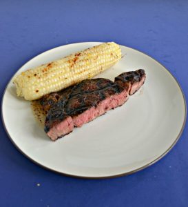 A plate with an ear of corn on the cob with a large piece of steak in front of it that hass grill marks on the top and a pink center with a blue background.