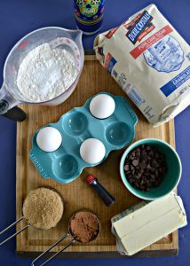 A cutting board topped with a measuring cup of flour, Dixie Crystals sugar in a bag, a blue container with 3 eggs in it, a bowl of chocolate chips, 2 sticks of butter, a measuring cup of brown sugar, and a cup of cocoa powder.