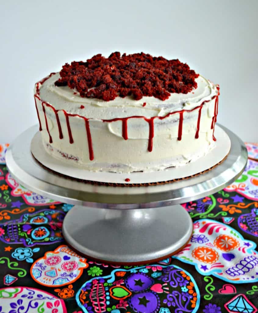A white cake sitting on a silver cake stand. The cake has red velvet crumbs on top and a red bloody drip around the edges. 