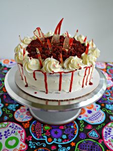 A silver cake stand with a white cake on top with a moun dof red velvet cake crumbs in the middle, bloody drizzle around the edges, and candy glass shards with a bloody drip sticking out of the cake.