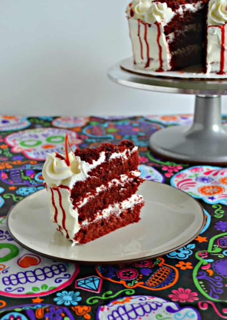 A large slice of red velvet cake with three layers in between buttercream frosting with a red drizzle running down the side and a candy glass shard sticking out of it. In the upper right hand corner you can see a silver cake stand with a white cake and bloody drizzle on it. 