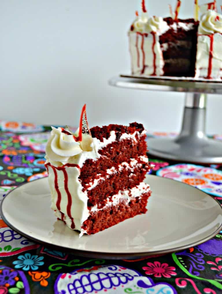 A large slice of red velvet cake with three layers in between buttercream frosting with a red drizzle running down the side and a candy glass shard sticking out of it. In the upper right hand corner you can see a silver cake stand with a white cake and bloody drizzle on it.