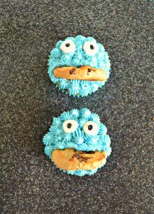 Two cupcakes positioned vertically and decorated with blue frosting, two candy eyes, and half a cookie mouth on a black background.