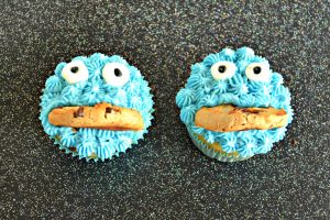 Two cupcakes decorated with blue frosting, two candy eyes, and half a cookie mouth on a black background.