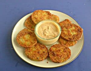 A side view of a plate piled with golden brown fried green tomatoes with a bowl of spicy remoulade in the middle on a blue background.