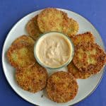 A plate piled with golden brown fried green tomatoes with a bowl of spicy remoulade in the middle on a blue background.