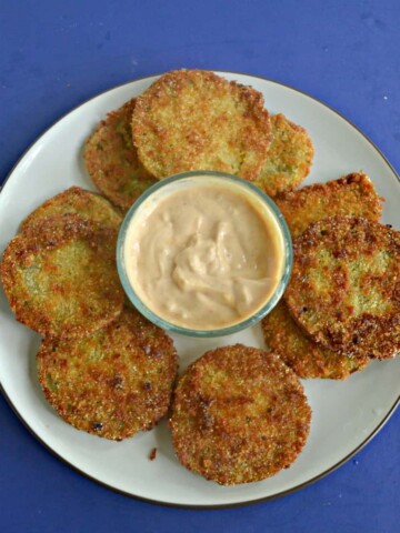 A plate piled with golden brown fried green tomatoes with a bowl of spicy remoulade in the middle on a blue background.
