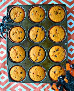 A muffin tin filled with orange cupcakes on an orange and white background with a spider coming onto the muffin tin from the bottom right and top left.
