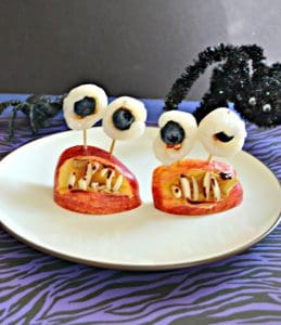 A plate with two monster mouths on it made of apple wedges, eyeballs made out of lychee with blueberries in them, and almond teeth, a spider creeping in from the back right, on a purple and black background.