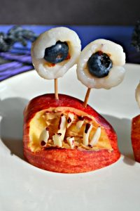 Close up image of a monster mouth made out of an apple wedge with almond sliver teeth, toothpicks coming out of the top spearing lychee and blueberry eyeballs.