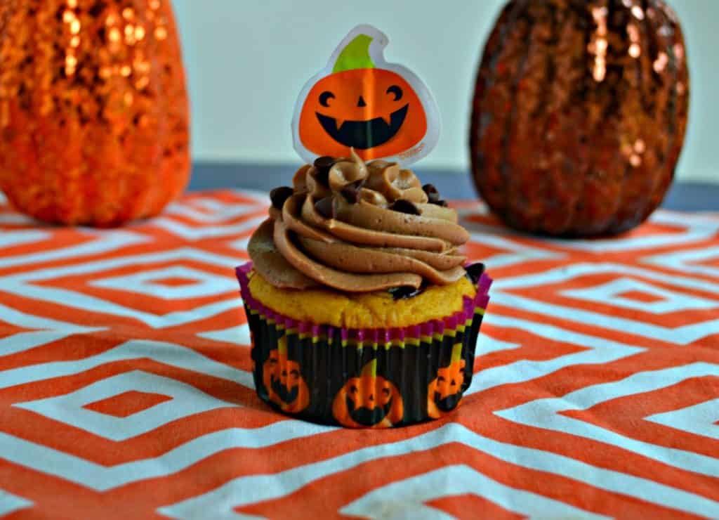 A close up view of a cupcake in a pumpkin cupcake liner topped with chocolate frosting, sprinkled with chocolate chips, topped with a paper pumpkin on an orange and white platemath. There are two pumpkin behind it, one on either side. 