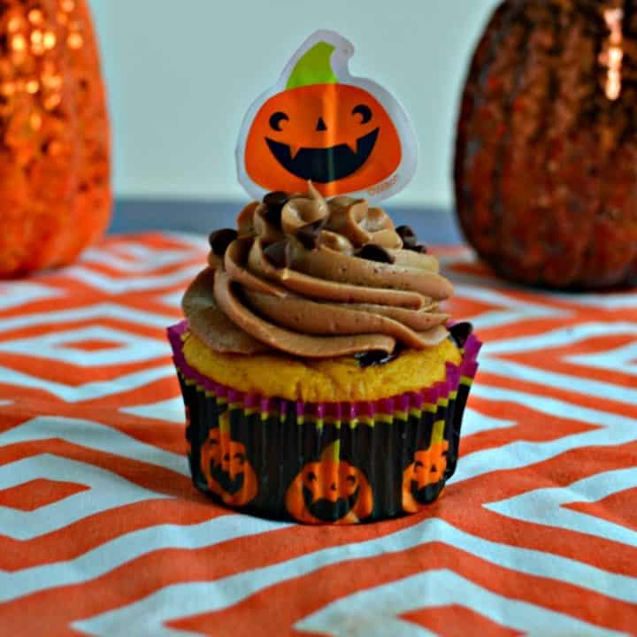 A close up view of a cupcake in a pumpkin cupcake liner topped with chocolate frosting, sprinkled with chocolate chips, topped with a paper pumpkin on an orange and white platemath. There are two pumpkin behind it, one on either side.