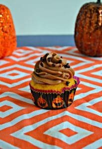 A close up view of a cupcake in a pumpkin cupcake liner topped with chocolate frosting, sprinkled with chocolate chips, on an orange and white platemat. There are two pumpkin behind it, one on either side.