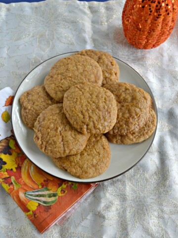 A white plate piled high with snickerdoodle cookies on top of an orange napkin with leaves on it and part of a pumpkin peeking in from the upper right hand corner.