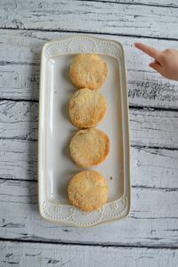 A white plate with 4 biscuits lined up on it on a white wooden backdrop with my toddler's finger pointing at the top biscuit.