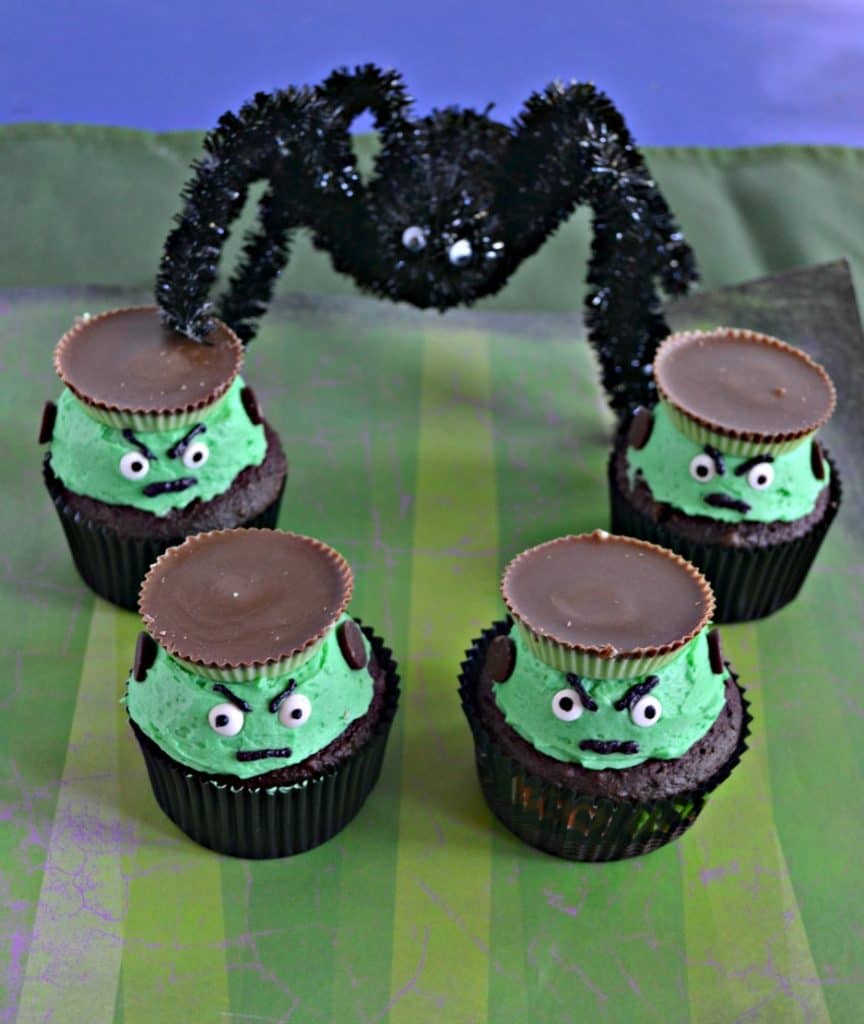 Four Frankenstein Cupcakes with green frosting, edible eyes, and a peanut butter cup hat sitting on a green striped background with a huge spider towering over them in the background.