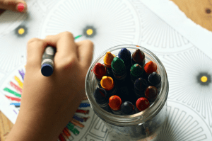 A top view of a cup filled with crayons and on the left a child's hand coloring with a green crayon.